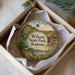A decorative ornament in a white box, featuring a scenic landscape with a river, mountains, and a small building.