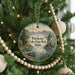 A round ornament hanging on a Christmas tree, featuring a scenic landscape with mountains, a lake, and a dock.