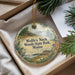 A round ornament with a scenic landscape of woods and a bird is displayed in a box, surrounded by pine branches.