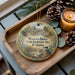 A wooden tray with a decorative ornament featuring a bird and a scenic landscape, along with pine cones and a lit candle.