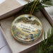 A decorative ornament in a box, featuring a bird and a scenic landscape with a bridge.