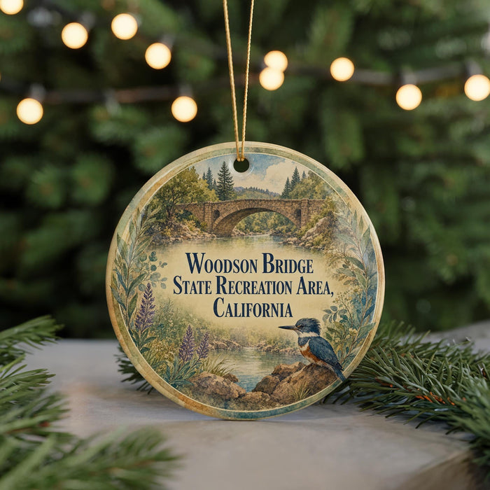 A circular ornament with a scenic landscape featuring a bridge, trees, and a bird, hanging in front of a Christmas tree with twinkling lights.