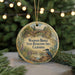 A circular ornament with a scenic landscape featuring a bridge, trees, and a bird, hanging in front of a Christmas tree with twinkling lights.