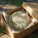 A round ornament in a wooden box, featuring a scenic landscape with a bridge and a bird, is displayed on a wooden surface.