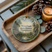 A decorative ornament featuring a scenic forest landscape with a river, pine trees, and a sign that reads "Wrentham State Forest, Massachusetts". The ornament is placed on a wooden tray, with pine cones and a lit candle nearby.