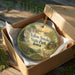 A decorative ornament in a wooden box, featuring a scenic landscape with a deer and a sunset.