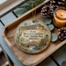 A decorative ornament featuring a cactus and a bridge, with the text "Yuma Quartermaster Depot State Historic Park, Arizona" displayed on it. The ornament is placed on a wooden tray, with pine cones and a lit candle nearby.