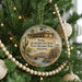 A round ornament hanging on a Christmas tree, featuring an illustration of a historic building and a desert landscape.