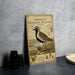 A vintage-style metal sign depicting an American golden-plover bird, with additional text and illustrations, is displayed on a gray countertop next to a stack of black camera lenses.