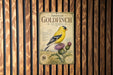 A vintage-style metal sign with an illustration of a yellow finch perched on a thistle plant, mounted on a wooden wall.