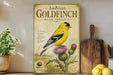 a vintage-style poster of an American Goldfinch on a thistle, displayed on a shelf next to a wooden cutting board and some potted plants.