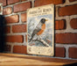 A vintage-style metal sign featuring an illustration of a robin perched on a branch with snow and pine trees in the background, mounted on a brick wall.