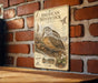a vintage-style metal plaque with an illustration of a woodcock bird, mounted on a brick wall.