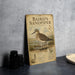 A metal sign with an illustration of a sandpiper bird, placed on a table next to some black camera lenses.