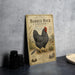 a vintage-style metal sign with an illustration of a black and white chicken, placed on a table next to some black camera lenses.