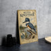 A metal sign featuring an illustration of a belted kingfisher, a type of wading bird, standing on a branch with a river and forest in the background.