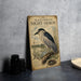 a vintage-style poster of a black-crowned night-heron, with a detailed illustration of the bird perched on a branch and a nest nearby. The poster is placed on a gray wall, and there are some black camera lenses visible in the foreground.