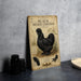 a metal sign with an illustration of a black Silkie chicken, placed on a table next to some black camera lenses.