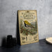 a vintage-style metal sign with a black-throated green warbler illustration, placed on a gray countertop next to some black camera lenses.