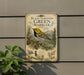 A vintage-style metal sign with a black-throated green warbler illustration, mounted on a gray wall next to a plant.