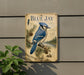 A vintage-style metal sign featuring a blue jay bird, pine cones, and pine needles, mounted on a wall.