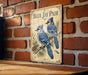 A vintage-style metal sign featuring illustrations of two blue jays perched on a branch, mounted on a brick wall.