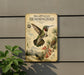 a vintage-style illustration of a hummingbird, with detailed illustrations of the bird's colorful feathers, wings, and beak. The hummingbird is surrounded by various plants and flowers, creating a lush and vibrant scene. The image is mounted on a wall, and there is a plant visible in the foreground.
