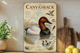a vintage-style metal sign with a painting of a duck swimming in a pond, surrounded by various bird illustrations. The sign is titled "Canvasback" and includes text and illustrations related to the bird.