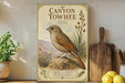 a vintage-style poster of a bird, specifically a Towhee, perched on a rock in a desert setting. The poster is displayed on a shelf next to a wooden cutting board and a potted plant.