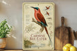 A vintage-style metal sign featuring a colorful illustration of a bird, specifically a carmine bee-eater, perched on a branch. The sign is surrounded by various kitchen utensils and decor items, such as a wooden cutting board, a potted plant, and a bowl of fruit.