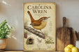 a poster of Carolina Wren, a bird, displayed on a shelf next to a wooden cutting board and some potted plants.