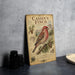 a vintage-style metal plaque with an illustration of a red bird perched on a branch, surrounded by pine cones and other natural elements.