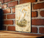 a metal sign with an illustration of a white bird, likely a heron or egret, standing on a grassy field. The sign is mounted on a brick wall, and there is a wooden table below it.