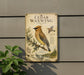 a vintage-style metal sign with a bird illustration, mounted on a wall next to a plant.