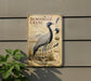 a vintage-style metal plate with an illustration of a demoiselle crane, a tall bird with a black and white plumage. The plate is mounted on a wall, and there is a plant visible to the left of the plate.