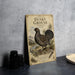 A metal sign with an illustration of a grouse bird, labeled as a "Dusky grouse", is displayed on a table next to some black camera lenses.