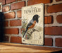 a vintage-style sign with a black and white illustration of a bird, likely a Towhee, perched on a branch. The sign is mounted on a brick wall, and there is a wooden surface below it.