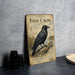 a vintage-style metal sign with a black crow illustration, placed on a gray countertop next to a stack of black camera lenses.