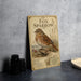 a vintage-style metal sign with a detailed illustration of a brown and white bird, likely a fox sparrow, standing on a branch with berries. The sign is placed on a gray countertop next to a stack of black camera lenses and a white dish.