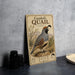 A vintage-style metal sign featuring an illustration of a quail bird, with text and decorative elements, placed on a gray countertop next to some black camera lenses.