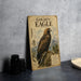 a vintage-style metal sign with an illustration of a golden eagle, placed on a countertop next to some camera equipment.