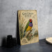 A vintage-style metal sign with a colorful illustration of a Gouldian finch, featuring text and a map, is displayed on a gray countertop next to some black camera lenses.