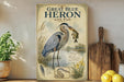 A framed illustration of a great blue heron with a fish in its beak, standing in a body of water surrounded by plants and flowers.