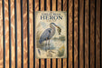 A vintage-style sign featuring a great blue heron with a fish in its beak, set against a wooden background.