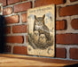 a vintage-style poster of a great horned owl with its young nestled in a tree, mounted on a brick wall.