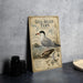 a vintage-style metal plate with a detailed illustration of a gull-billed tern, a type of seabird, perched on a rock. The plate is placed on a table next to a stack of black camera lenses and a woven basket.