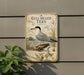a vintage-style metal sign with a detailed illustration of a gull-billed tern, a type of seabird, perched on a rock. The sign is mounted on a wall and is surrounded by greenery, including a plant to the left.