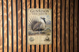 A metal sign with a picture of a bird, labeled "Gunnison Sage-Grouse", is mounted on a wooden wall.