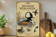 a poster of a hooded merican, a bird with a distinctive black and white head, sitting in a body of water surrounded by pine trees.