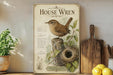 a framed illustration of a house wren, a small bird, perched on a tree stump with a nest nearby. The frame has a vintage-style design with decorative elements. The background of the image is a white wall, and there are some potted plants and kitchen utensils visible in the foreground.